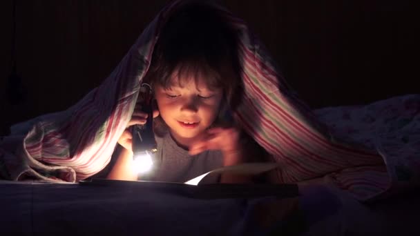 Niño leyendo un libro debajo de las cubiertas — Vídeo de stock