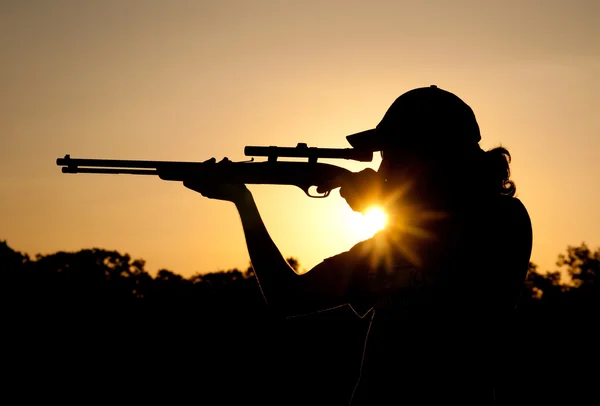 Silueta de un joven disparando con un rifle largo contra el cielo del atardecer, con un rayo de sol —  Fotos de Stock