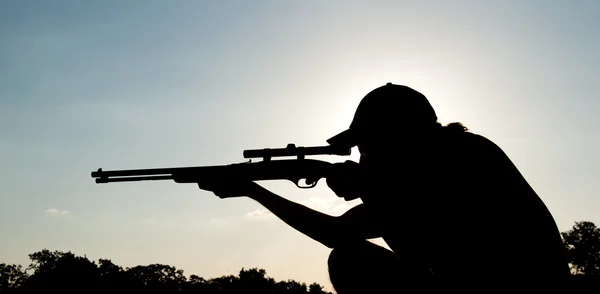 Silueta de un joven apuntando con un rifle largo contra la puesta del sol — Foto de Stock