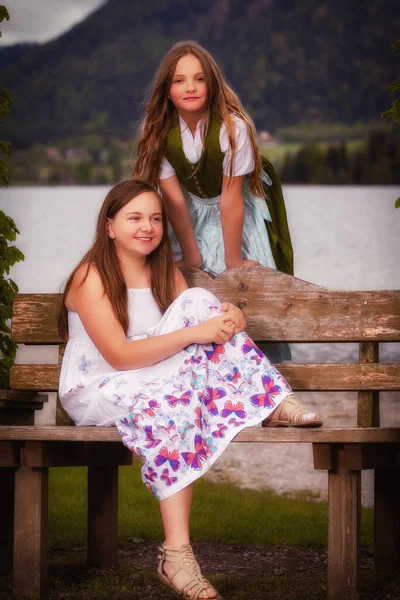 Две Сестры Длинными Волосами Летнем Платье Грязнуля Скамейке Перед Озером — стоковое фото