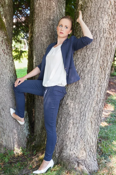 Jonge vrouw in blauwe jas leunend tegen bomen — Stockfoto