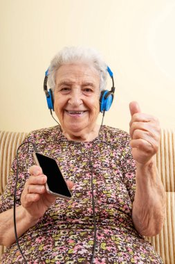 müzik dinleyen, sevimli yaşlı kadın evde akıllı telefondan veya cihazdan mavi kulaklıklarla müzik dinleyen. Ne dinlediğini göstermek için başparmağını kaldır. Yaşlı bir kadın elleriyle imza atabilir mi?