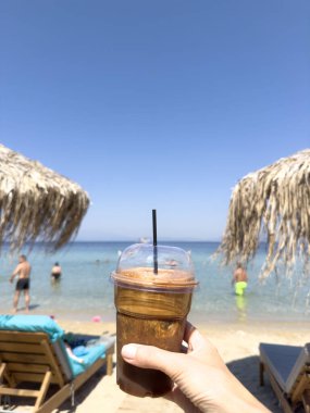 Tropik plajda deniz ve gökyüzü manzaralı buzlu Yunan frappe kahvesi. Soğuk yaz tatili içeceği, soğuk kahve ve mavi gökyüzünün altında fotokopi alanı olan saman şemsiyeli deniz kenarı anı..