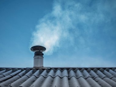 Paslanmaz çelik baca, tahta çatıda. Mavi gökyüzüne doğru duman yayıyor.