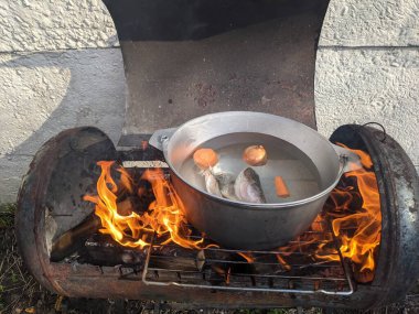 Kırmızı balık çorbasının baharında ateşte pişirmek. Gölün yanındaki ateşte kulağı olan siyah melon şapka. Tahtadan yapılmış bir çorba yalağı. Ateşin üstünde lezzetli balık çorbası