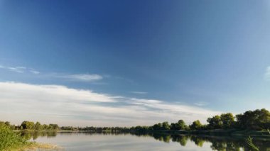 Sakin göl manzarası sakin su, mavi gökyüzü ve yansımalarla dolu. Çerçeveli sazlıklar, ağaçlar, 