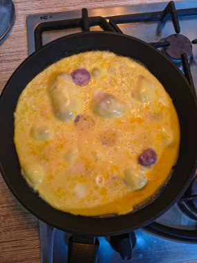 Dilimlenmiş sosisli omlet, gaz sobasında siyah bir tavada pişiyor. Sıcak ışıklandırma, görünür doku ve mutfak ayrıntıları rahat bir kahvaltı sahnesi yaratır..