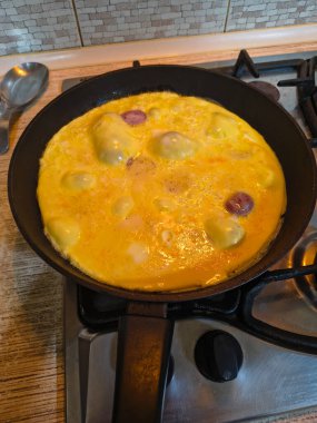 Dilimlenmiş sosisli omlet, gaz sobasında siyah bir tavada pişiyor. Sıcak ışıklandırma, görünür doku ve mutfak ayrıntıları rahat bir kahvaltı sahnesi yaratır..