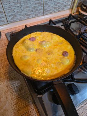 Dilimlenmiş sosisli omlet, gaz sobasında siyah bir tavada pişiyor. Sıcak ışıklandırma, görünür doku ve mutfak ayrıntıları rahat bir kahvaltı sahnesi yaratır..