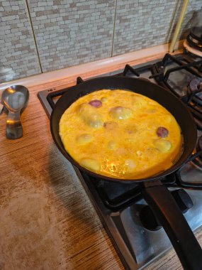 Dilimlenmiş sosisli omlet, gaz sobasında siyah bir tavada pişiyor. Sıcak ışıklandırma, görünür doku ve mutfak ayrıntıları rahat bir kahvaltı sahnesi yaratır..