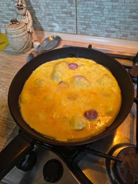 Dilimlenmiş sosisli omlet, gaz sobasında siyah bir tavada pişiyor. Sıcak ışıklandırma, görünür doku ve mutfak ayrıntıları rahat bir kahvaltı sahnesi yaratır..