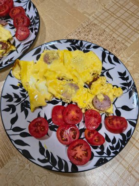 Çırpılmış yumurta, sosis dilimleri ve taze domates dilimleriyle kahvaltı tabağı. Bej desenli bir masa örtüsünün üzerinde dekoratif yaprak desenli bir tabakta servis edilmiş..