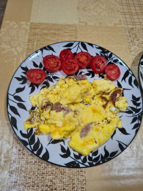 Çırpılmış yumurta, sosis dilimleri ve taze domates dilimleriyle kahvaltı tabağı. Bej desenli bir masa örtüsünün üzerinde dekoratif yaprak desenli bir tabakta servis edilmiş..