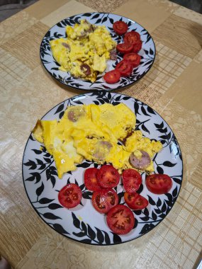 Çırpılmış yumurta, sosis dilimleri ve taze domates dilimleriyle kahvaltı tabağı. Bej desenli bir masa örtüsünün üzerinde dekoratif yaprak desenli bir tabakta servis edilmiş..