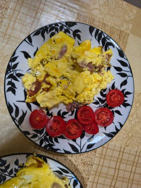 Çırpılmış yumurta, sosis dilimleri ve taze domates dilimleriyle kahvaltı tabağı. Bej desenli bir masa örtüsünün üzerinde dekoratif yaprak desenli bir tabakta servis edilmiş..