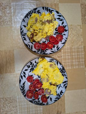 Çırpılmış yumurta, sosis dilimleri ve taze domates dilimleriyle kahvaltı tabağı. Bej desenli bir masa örtüsünün üzerinde dekoratif yaprak desenli bir tabakta servis edilmiş..