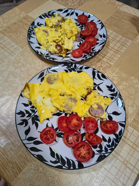 Çırpılmış yumurta, sosis dilimleri ve taze domates dilimleriyle kahvaltı tabağı. Bej desenli bir masa örtüsünün üzerinde dekoratif yaprak desenli bir tabakta servis edilmiş..