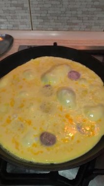 Dilimlenmiş sosisli omlet, gaz sobasında siyah bir tavada pişiyor. Sıcak ışıklandırma, görünür doku ve mutfak ayrıntıları rahat bir kahvaltı sahnesi yaratır..