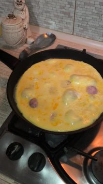 Dilimlenmiş sosisli omlet, gaz sobasında siyah bir tavada pişiyor. Sıcak ışıklandırma, görünür doku ve mutfak ayrıntıları rahat bir kahvaltı sahnesi yaratır..