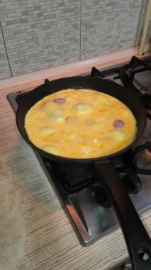 Dilimlenmiş sosisli omlet, gaz sobasında siyah bir tavada pişiyor. Sıcak ışıklandırma, görünür doku ve mutfak ayrıntıları rahat bir kahvaltı sahnesi yaratır..