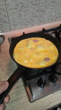 Dilimlenmiş sosisli omlet, gaz sobasında siyah bir tavada pişiyor. Sıcak ışıklandırma, görünür doku ve mutfak ayrıntıları rahat bir kahvaltı sahnesi yaratır..