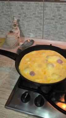 Dilimlenmiş sosisli omlet, gaz sobasında siyah bir tavada pişiyor. Sıcak ışıklandırma, görünür doku ve mutfak ayrıntıları rahat bir kahvaltı sahnesi yaratır..
