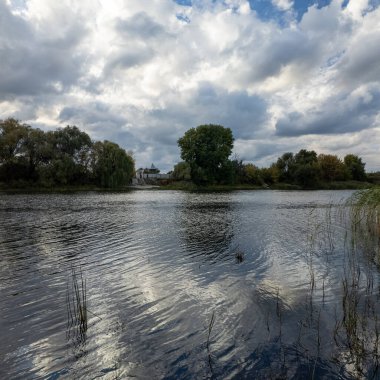 Sakin nehir ya da göl, dalgalı suyla çevrili, dramatik bulutlu gökyüzü altında sazlıklar ve ağaçlarla çevrili. Güneş ışığı bulutların arasından doğar, huzurlu, doğal bir sonbahar sahnesi yaratır..