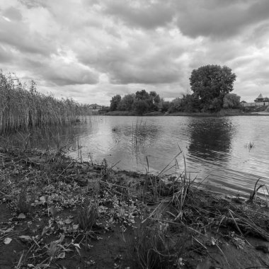 Sakin nehir ya da göl, dalgalı suyla çevrili, dramatik bulutlu gökyüzü altında sazlıklar ve ağaçlarla çevrili. Güneş ışığı bulutların arasından doğar, huzurlu, doğal bir sonbahar sahnesi yaratır..