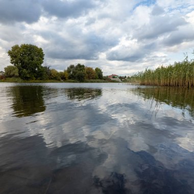 Sakin nehir ya da göl, dalgalı suyla çevrili, dramatik bulutlu gökyüzü altında sazlıklar ve ağaçlarla çevrili. Güneş ışığı bulutların arasından doğar, huzurlu, doğal bir sonbahar sahnesi yaratır..