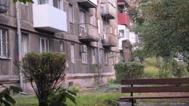 Çok katlı apartmanı, balkonları, ağaçları ve bankı olan şehir avlusu. Beton ve yeşillik karışımı, tipik bir Doğu Avrupa mahallesinde günlük yaşamı yakalıyor..