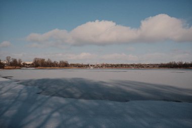 Donmuş göl manzarası, karla kaplı buzlar, sazlıklar ve mavi gökyüzü altındaki kış kıyı şeridi, sakin mevsimsel manzara soğuk havayı, durgun su kenarlarını ve kırsal kış atmosferini gösteriyor..