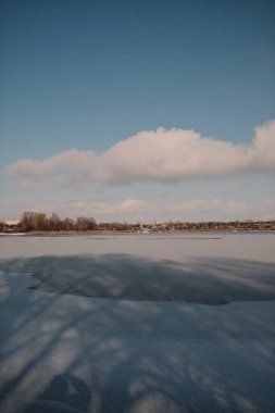 Donmuş göl manzarası, karla kaplı buzlar, sazlıklar ve mavi gökyüzü altındaki kış kıyı şeridi, sakin mevsimsel manzara soğuk havayı, durgun su kenarlarını ve kırsal kış atmosferini gösteriyor..