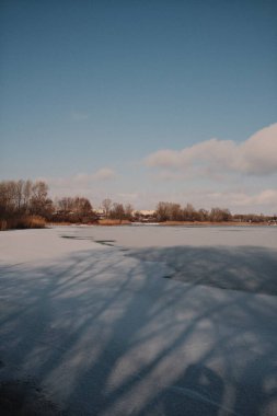 Donmuş göl manzarası, karla kaplı buzlar, sazlıklar ve mavi gökyüzü altındaki kış kıyı şeridi, sakin mevsimsel manzara soğuk havayı, durgun su kenarlarını ve kırsal kış atmosferini gösteriyor..