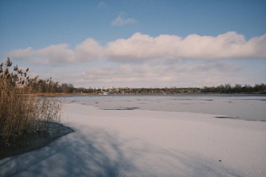 Donmuş göl manzarası, karla kaplı buzlar, sazlıklar ve mavi gökyüzü altındaki kış kıyı şeridi, sakin mevsimsel manzara soğuk havayı, durgun su kenarlarını ve kırsal kış atmosferini gösteriyor..