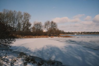 Donmuş göl manzarası, karla kaplı buzlar, sazlıklar ve mavi gökyüzü altındaki kış kıyı şeridi, sakin mevsimsel manzara soğuk havayı, durgun su kenarlarını ve kırsal kış atmosferini gösteriyor..