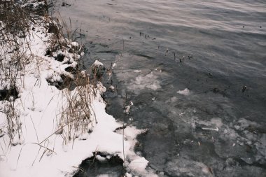 Buzlu göl kıyılarında eriyen karlar ve kıyıların yakınında kırılan buzlar soğuk dokuları, donmuş dalgaları ve sazlıkları gösteren kış geçiş sahneleri.,