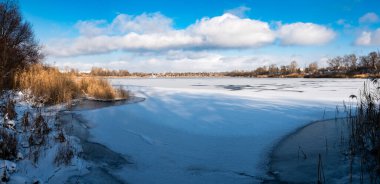 Parlak mavi gökyüzünün altında sakin bir kış donmuş gölü. Karla kaplı buz ayak izleri ve ağaç gölgeleri kıyıda altın kuru sazlıklar, çıplak ağaçlar ve soğuk güneş ışığı altında uzak ufuklar..