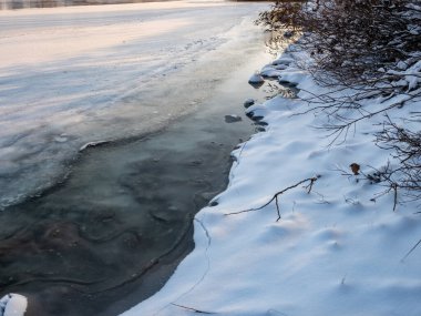 Parlak mavi gökyüzünün altında sakin bir kış donmuş gölü. Karla kaplı buz ayak izleri ve ağaç gölgeleri kıyıda altın kuru sazlıklar, çıplak ağaçlar ve soğuk güneş ışığı altında uzak ufuklar..