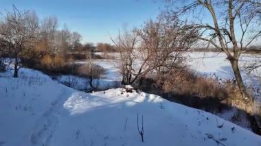 Parlak mavi gökyüzünün altında sakin bir kış donmuş gölü. Karla kaplı buz ayak izleri ve ağaç gölgeleri kıyıda altın kuru sazlıklar, çıplak ağaçlar ve soğuk güneş ışığı altında uzak ufuklar..