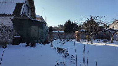 Kışın kırsal alanda gün batımında. Çıplak meyve ağaçları olan karla kaplı bahçe tavan arası olan eski tuğla ev, çit, açık mavi gökyüzü ve parlak alçak güneş. Soğuk, huzurlu kırsal atmosfer.