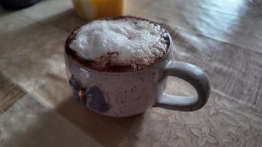 Mavi fiyonklu seramik bardakta köpüklü bir kapuçinonun samimi yakın çekimi. Yumuşak, iç mekan aydınlatması ahşap bir masada sıcak bir atmosfer yaratıyor, kızlık zarı teması için mükemmel..