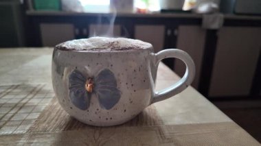 Mavi fiyonklu seramik bardakta köpüklü bir kapuçinonun samimi yakın çekimi. Yumuşak, iç mekan aydınlatması ahşap bir masada sıcak bir atmosfer yaratıyor, kızlık zarı teması için mükemmel..