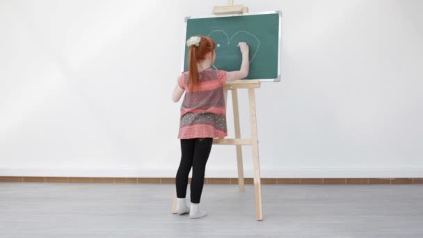 The little red-haired girl with freckles draws heart on the blackboard — Stock Video