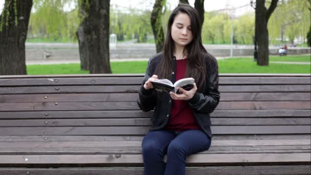 Женщина сидит на скамейке в парке и читает книгу — стоковое видео