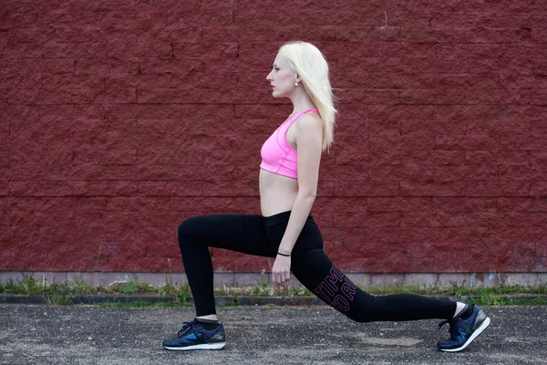 Young blonde woman is doing squat outdoors, red wall background — Stock Photo, Image