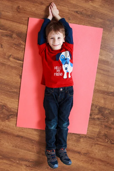 Niño en una camisa roja está acostado en una alfombra, haciendo ejercicio — Foto de Stock