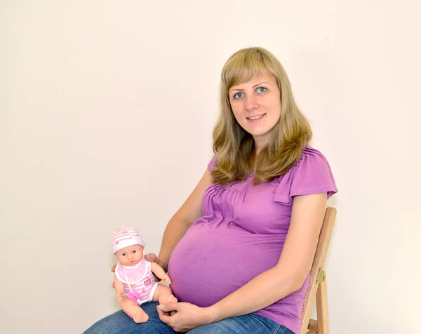Mladá těhotná žena sedí na židli s panenkou v rukou — Stock fotografie