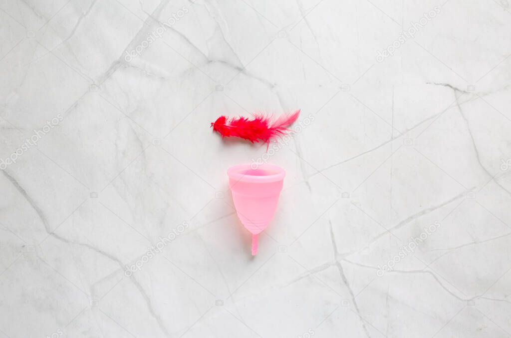 Menstrual cup and red bird feather on marble background. Alternative feminine hygiene product during the period. Women health concept