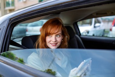 Akıllı Telefonlu Gülümseyen Kızıl Kadın Araba Penceresinden Bakıyor, Arka Koltukta Mutlu Kadın Yolcu, Yolculuk Paylaşımı ve Modern Ulaşım Konsepti, Şehir Seyahatinden Hoşlanan Genç Kız.