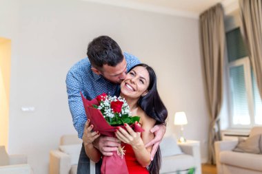 Sevecen bir adam bir buket kırmızı gül sunarken gülümseyen kız arkadaşını öpüyor. Romantik genç çift Sevgililer Günü 'nü kutluyor ya da parlak bir oturma odasında nişan sürprizi yapıyor..